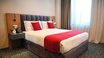 Una habitación de hotel con una cama grande, un cabecero de madera, una mesita de noche con lámpara y una ventana con cortinas.