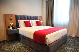 Una habitación de hotel con una cama grande, un cabecero de madera, una mesita de noche con lámpara y una ventana con cortinas.