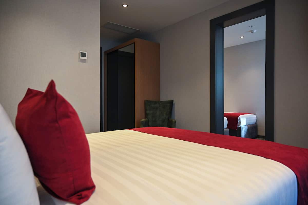 A hotel room with a bed, a red and white pillow, a chair, and a small table with a red cloth.