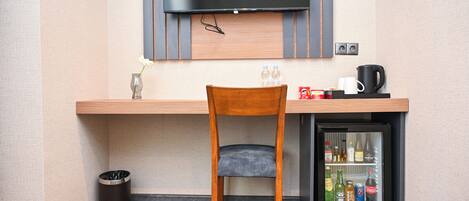 A compact hotel room with a small desk, a chair, a television mounted above the desk, a mini-fridge with beverages, and a trash bin.