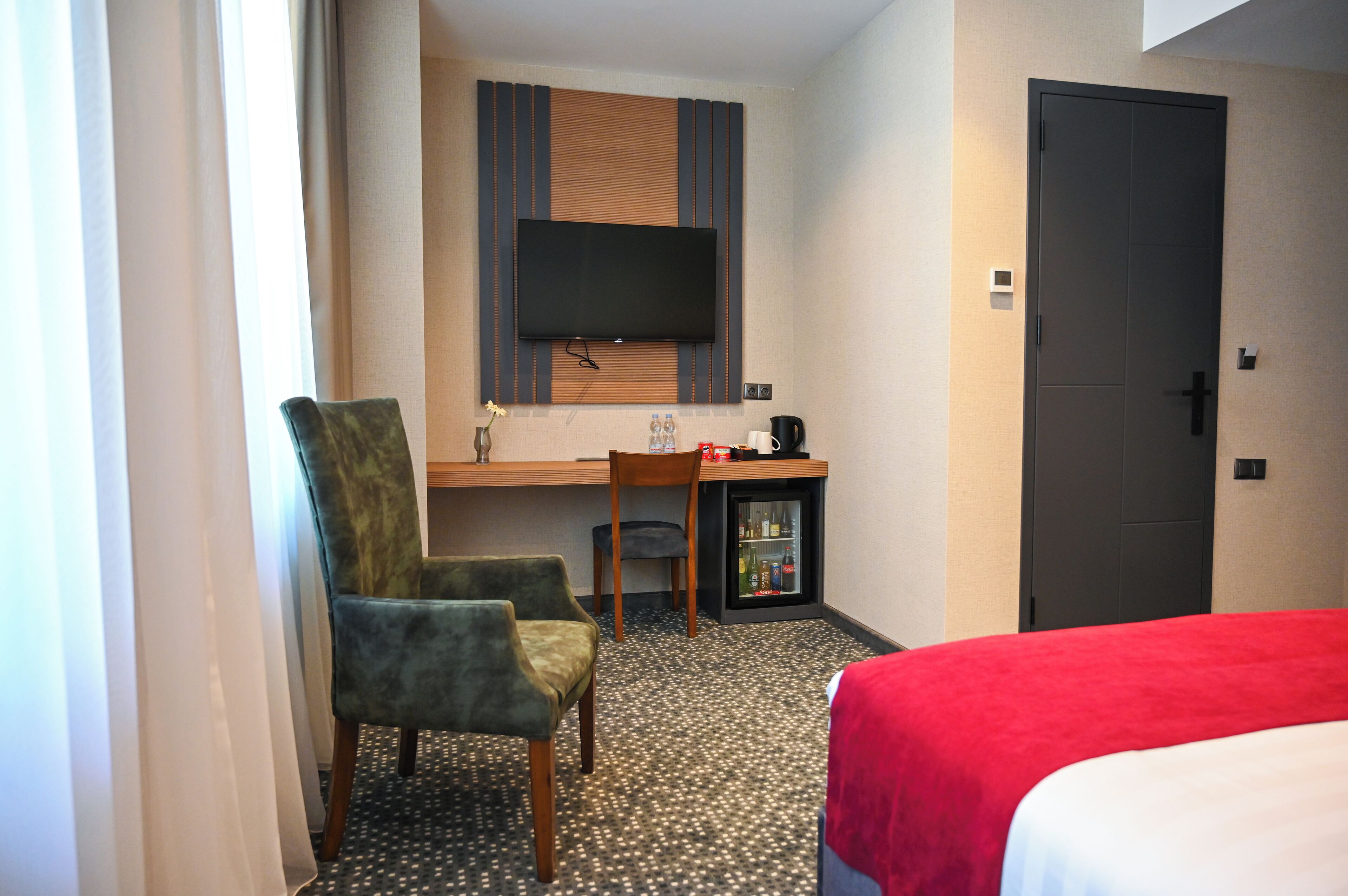 Ein Hotelzimmer mit Bett, Schreibtisch mit Fernseher, Sessel, Minibar und Tür.