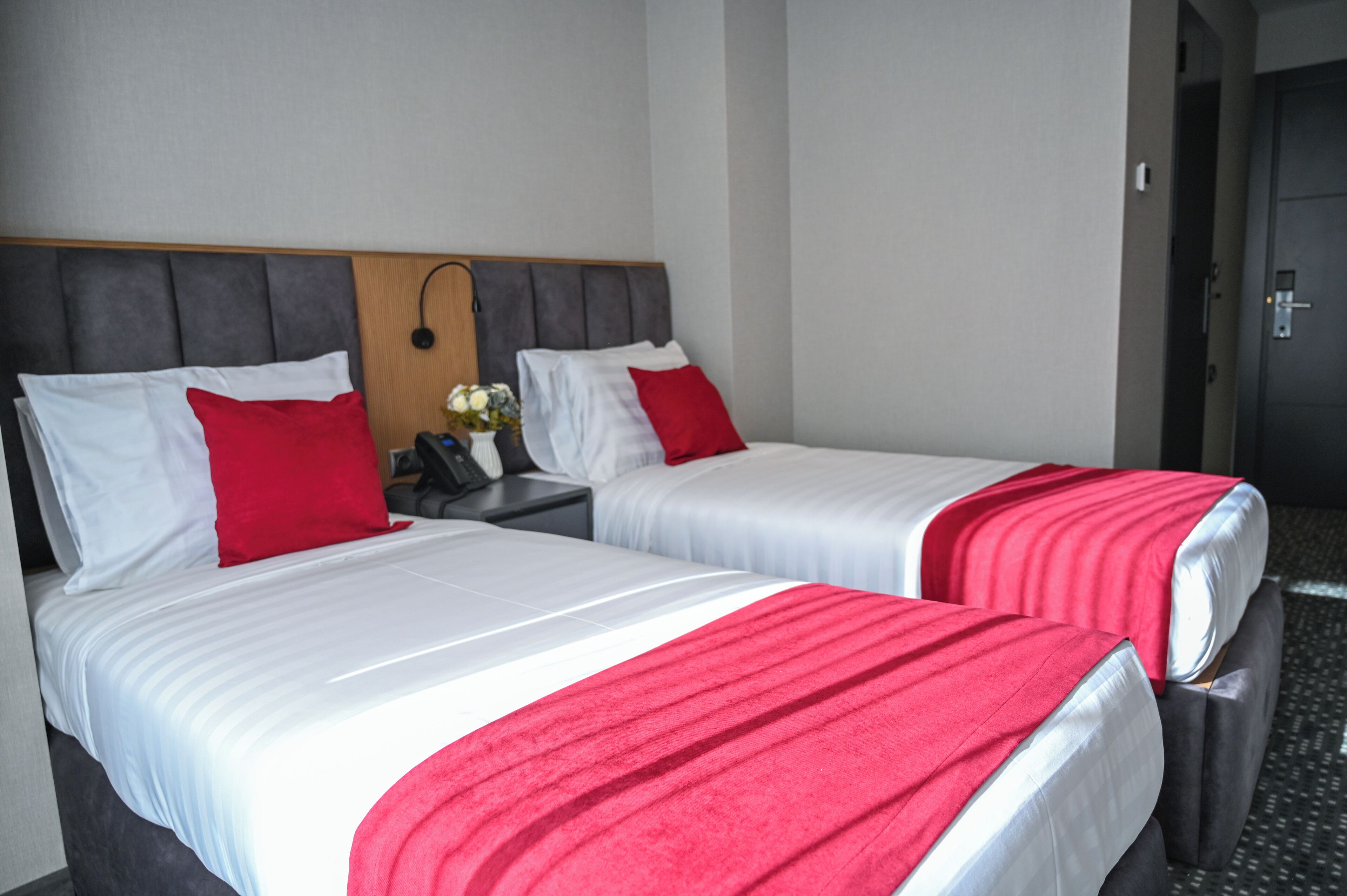Two beds with white and red bedding, a wooden headboard, a nightstand with a phone and vase, and a door in the background.