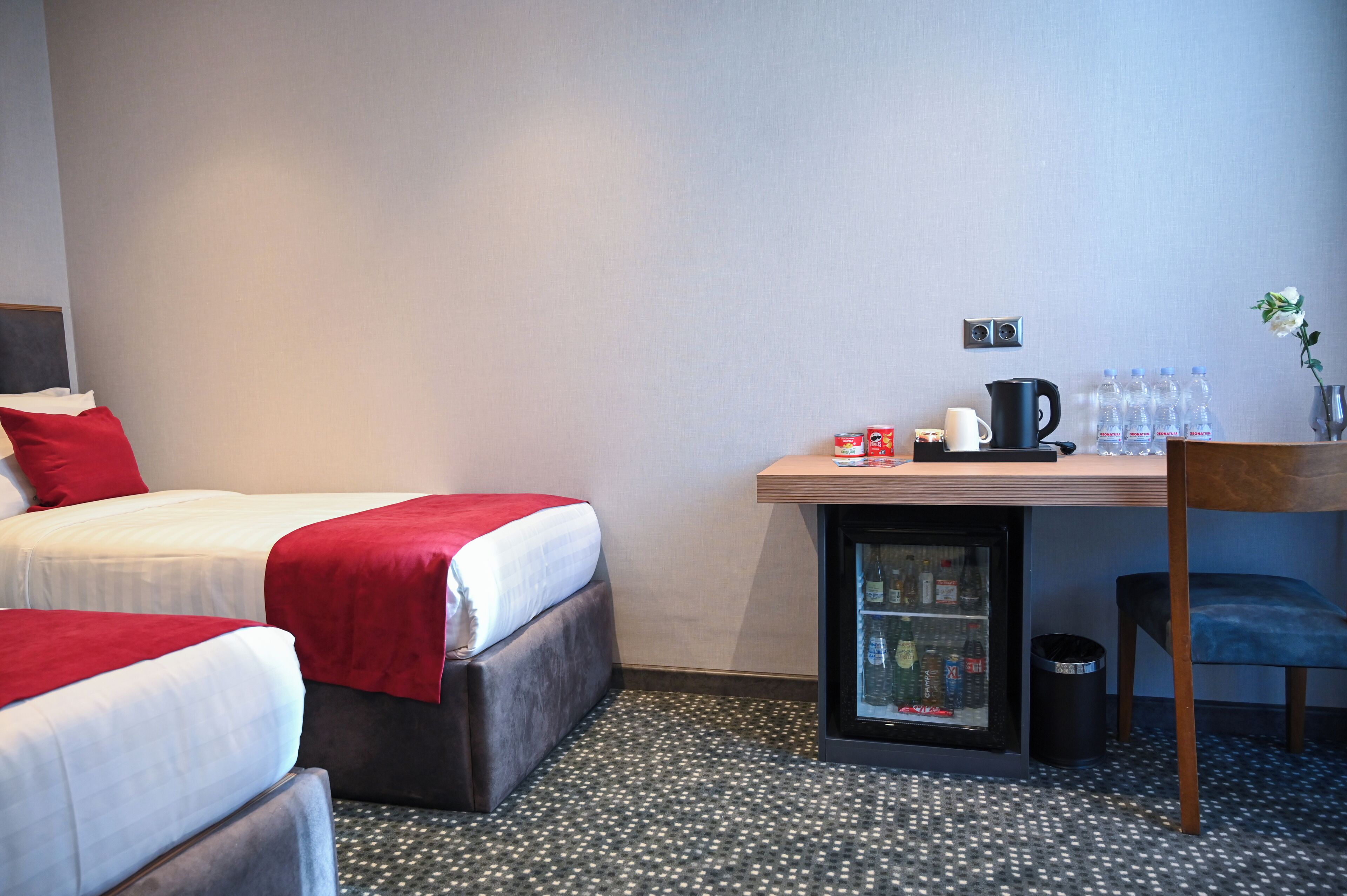 A hotel room with two beds, a small table with a kettle, cups, and bottles, a trash bin, and a refrigerator.