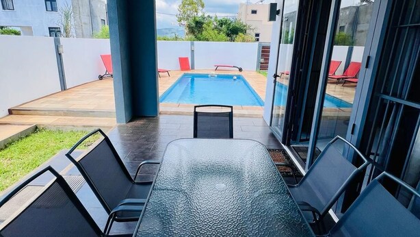 Ein überdachter Patio-Bereich mit Glastisch und Stühlen, ein Pool im Hintergrund und Blick auf benachbarte Gebäude.