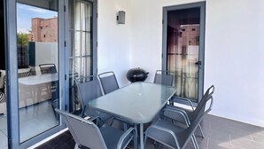 Ein moderner Essbereich im Freien mit einem Glastisch, Metallstühlen, großen Schiebegläsern und Blick auf ein Gebäude im Hintergrund.