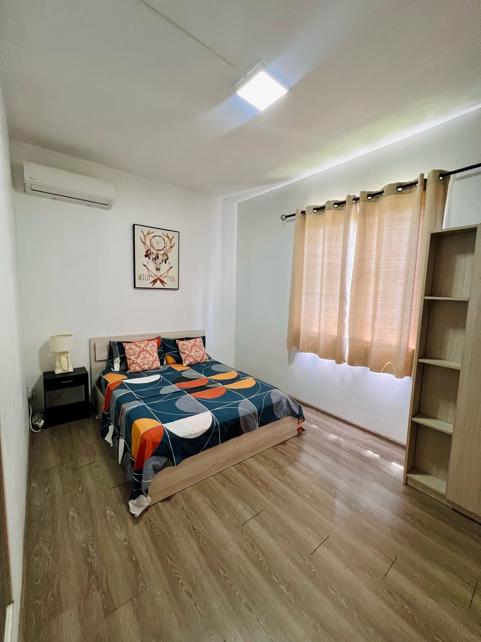 Een slaapkamer met een bed, een nachtkastje, een wandairconditioner, een raam met beige gordijnen en een houten vloer.