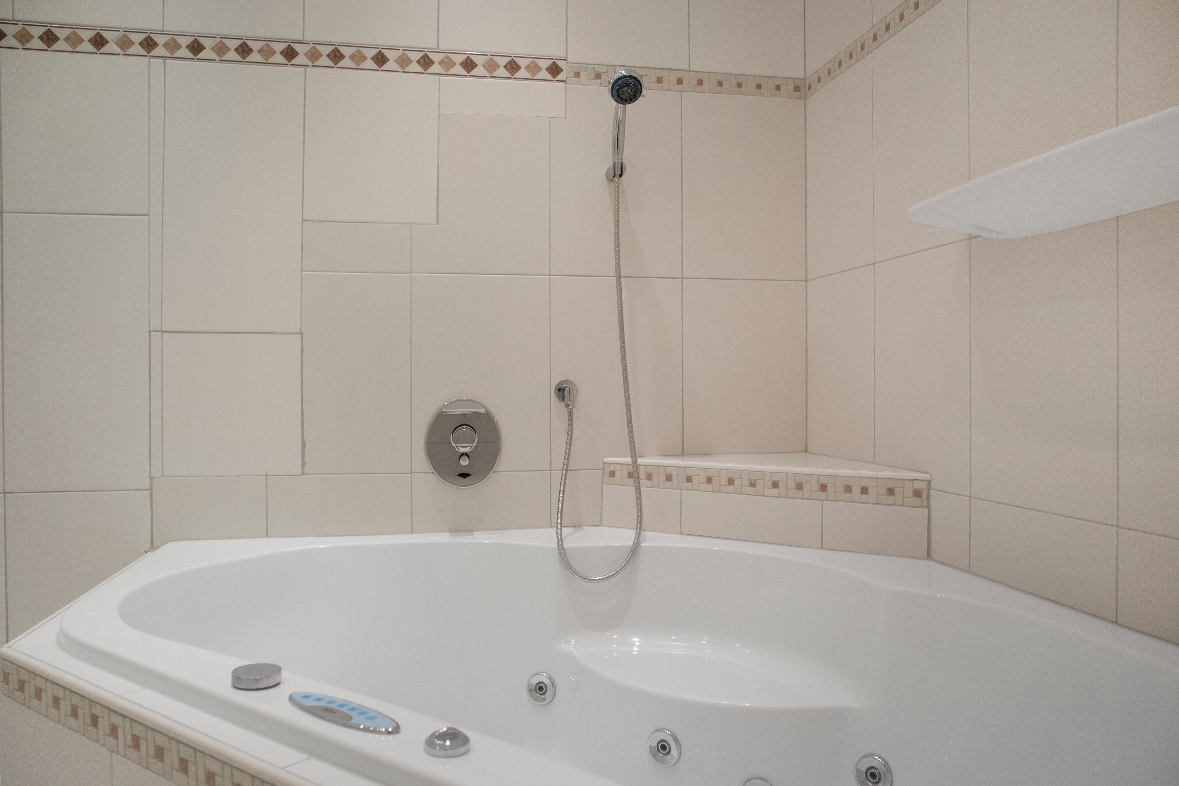 Une baignoire blanche dotée d’un pommeau de douche à main et d’un panneau de commande, le tout installé sur des murs carrelés agrémentés d’une bordure décorative.