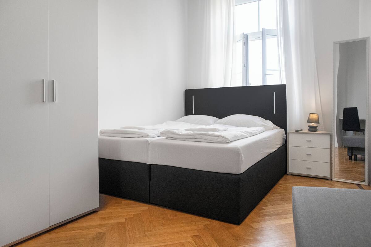 Ein modernes Schlafzimmer mit einem Bett mit schwarzem Kopfteil, weißer Bettwäsche, Holzboden, einem weißen Kleiderschrank, einem Nachttisch mit Lampe und einem Spiegel.