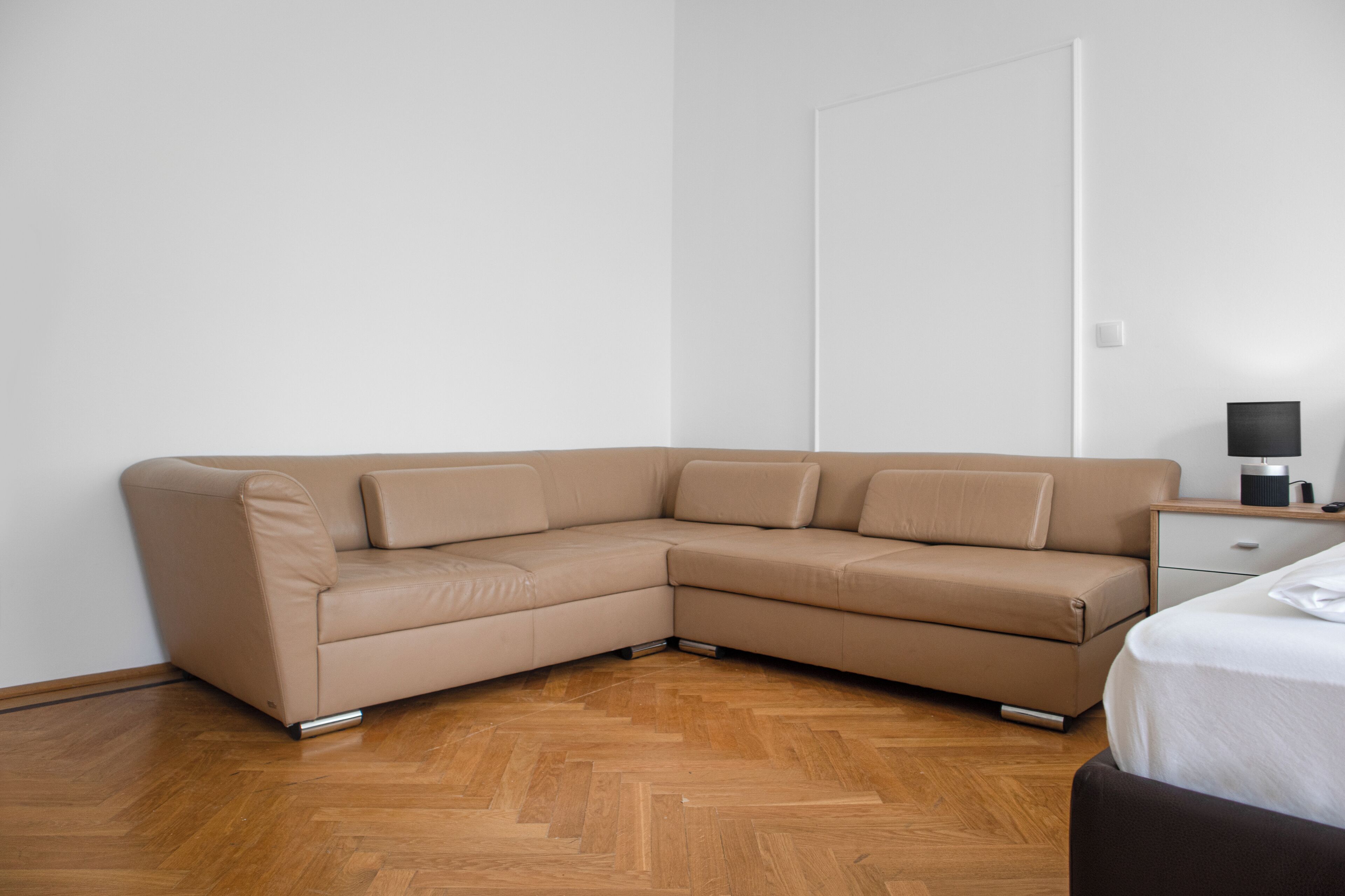 Un salon d’angle avec un canapé d’angle beige, un parquet en bois à motifs chevrons, un mur blanc et une petite table de chevet avec une lampe.