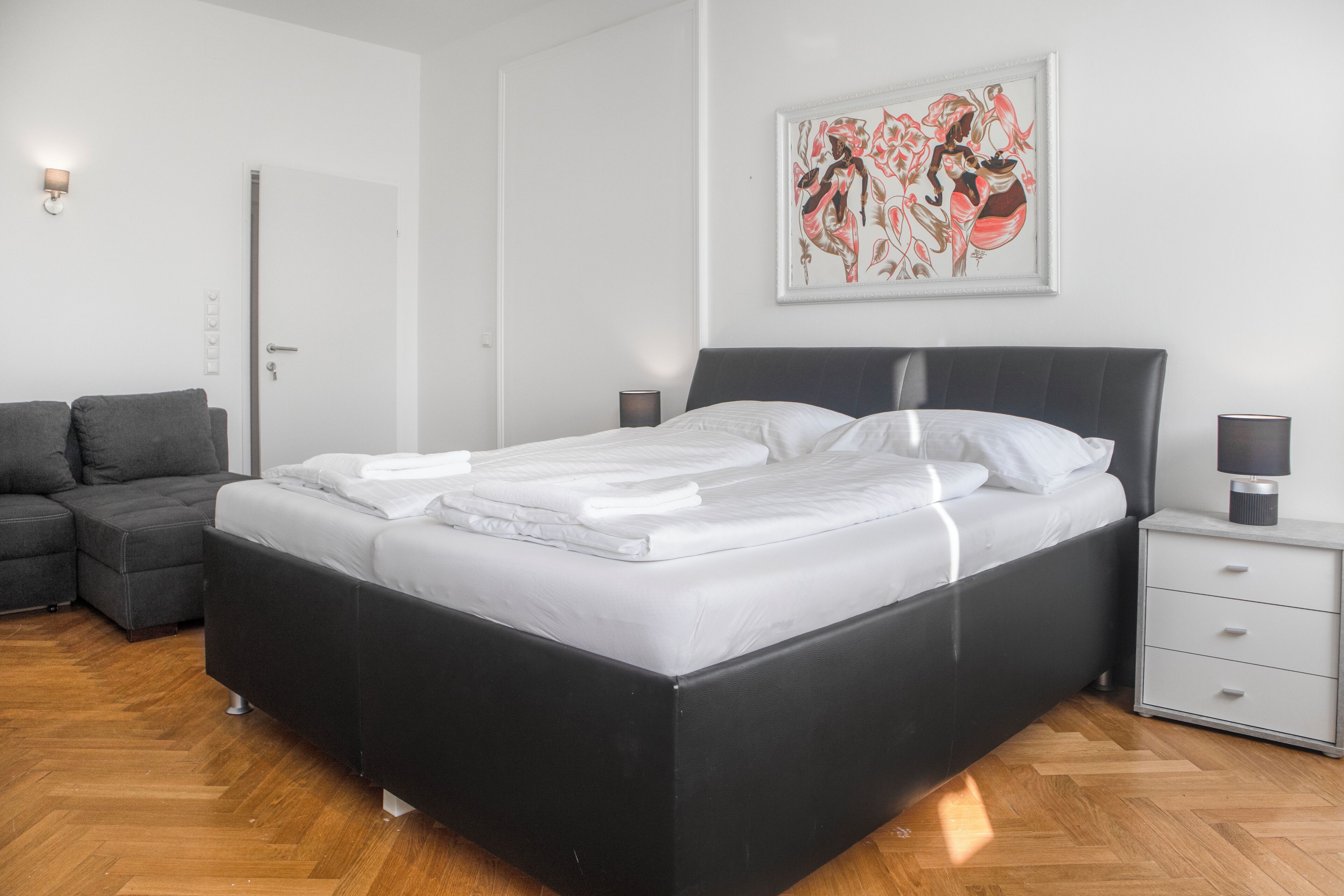 A modern bedroom with a black bed, white bedding, a grey sofa, a nightstand with a lamp, and a framed artwork on the wall.