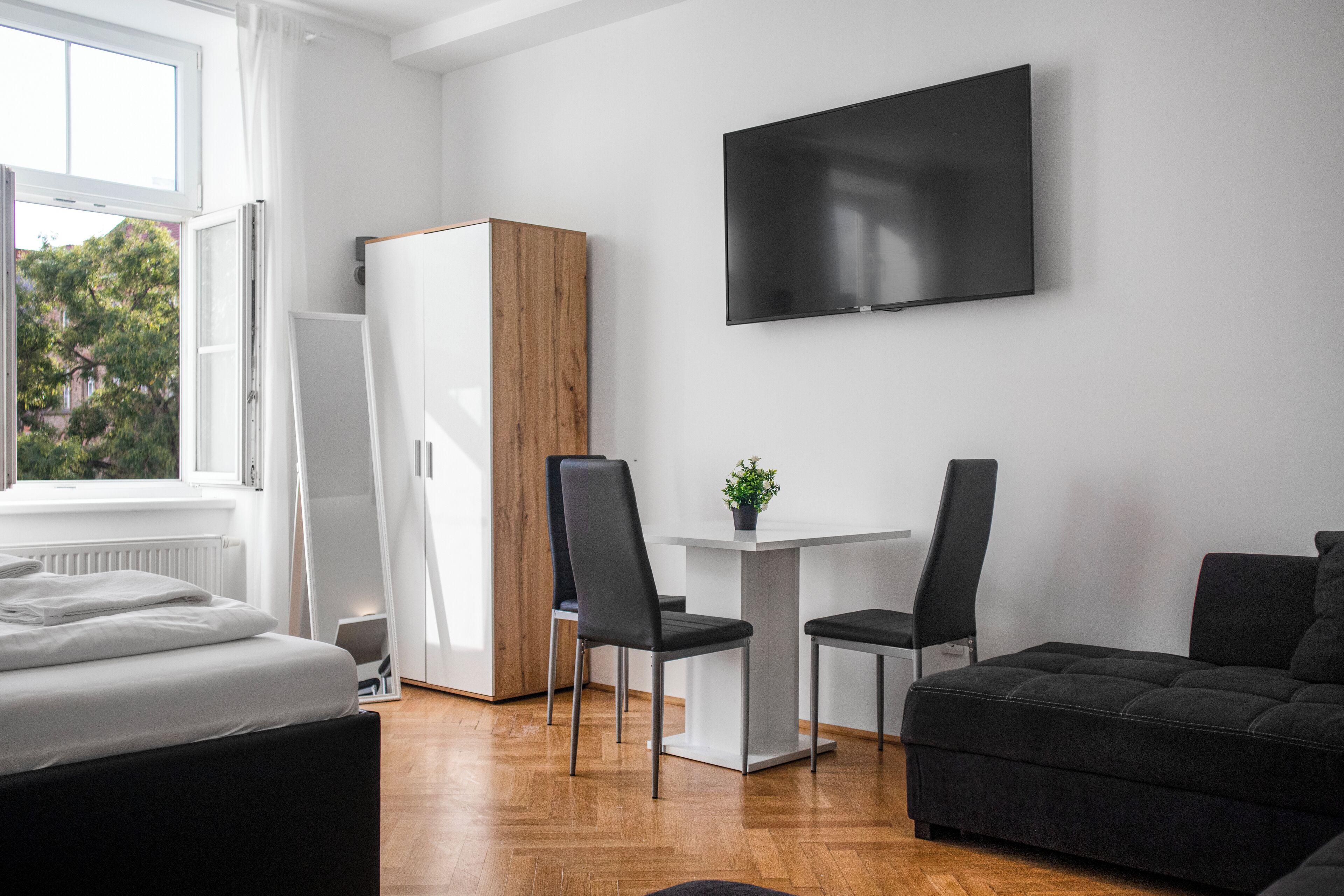 A modern, well-lit room with a bed, a sofa, a dining table with chairs, a TV mounted on the wall, and a wardrobe.