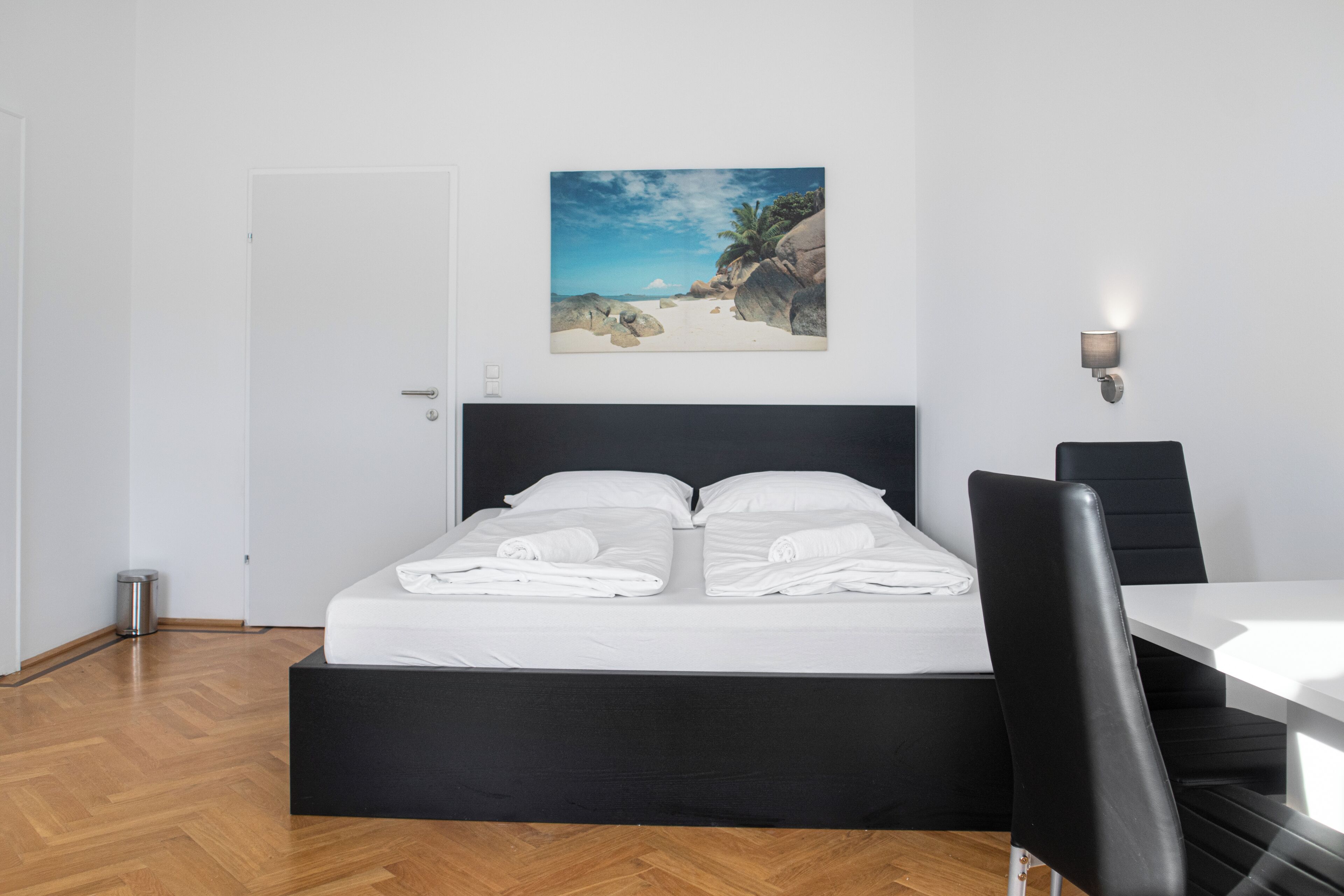 A neatly made bed with white linens, a black headboard, and a painting of a beach scene above it.