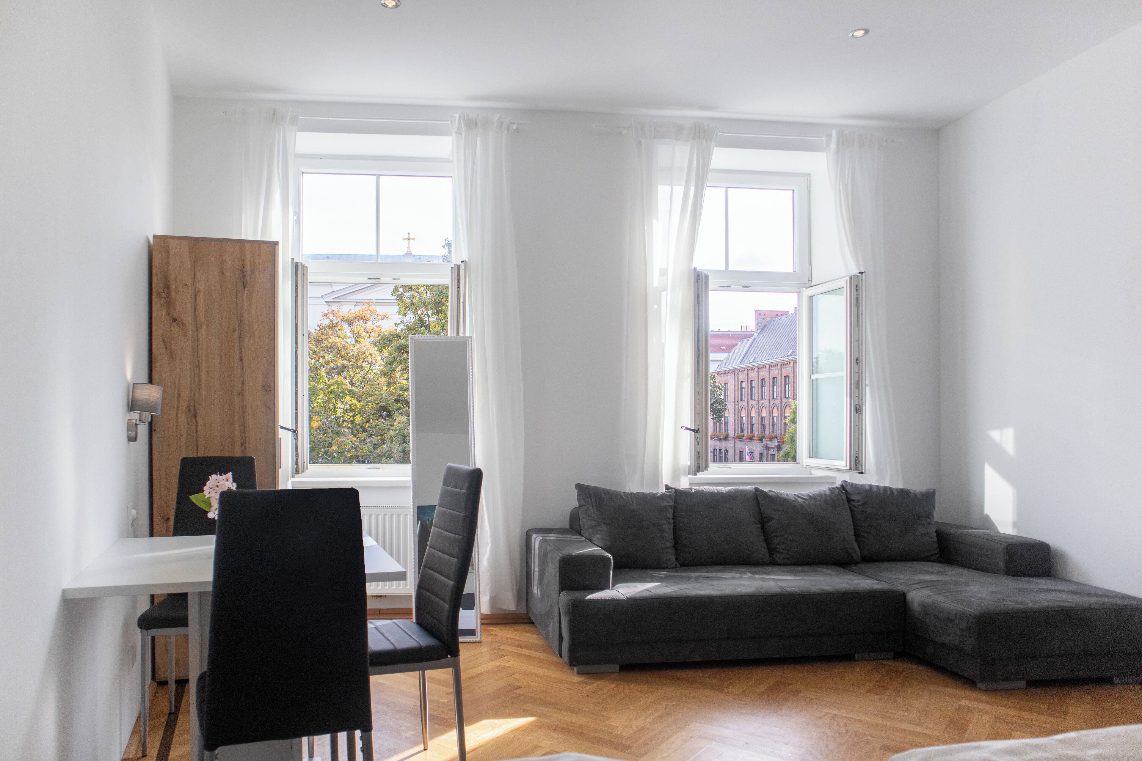 A modern living room with a grey sectional sofa, a dining table with chairs, and large windows with white curtains.