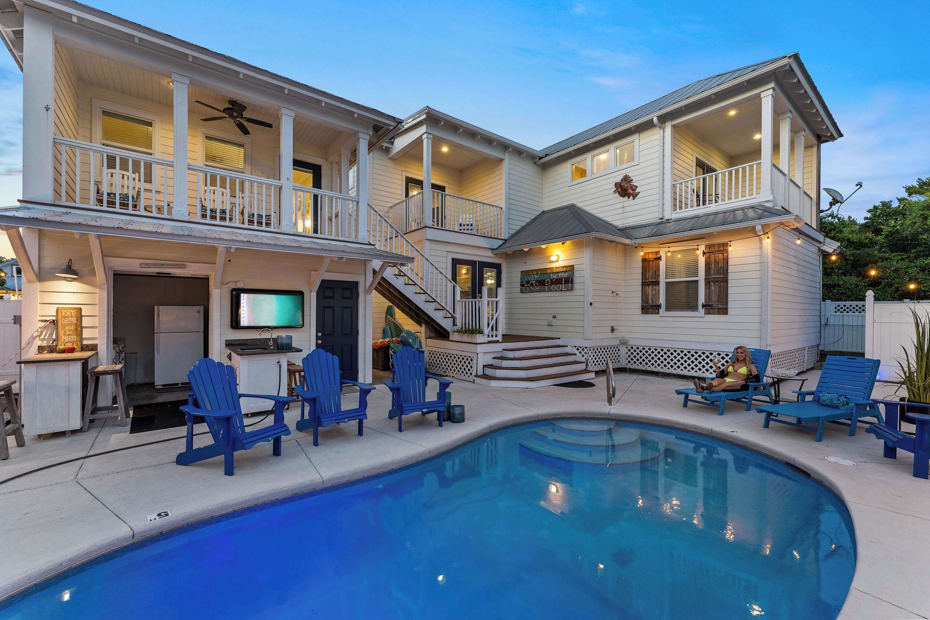 Une maison à deux étages avec une piscine, un espace salon extérieur et un panneau indiquant l’emplacement d’un bar au bord de la piscine.
