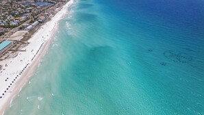 An aerial view of a beach with clear turquoise waters, a long stretch of sandy beach, and a row of beach umbrellas. - Lighthouse Legends-Free Golf Cart-1 Block to Beach (Destin)