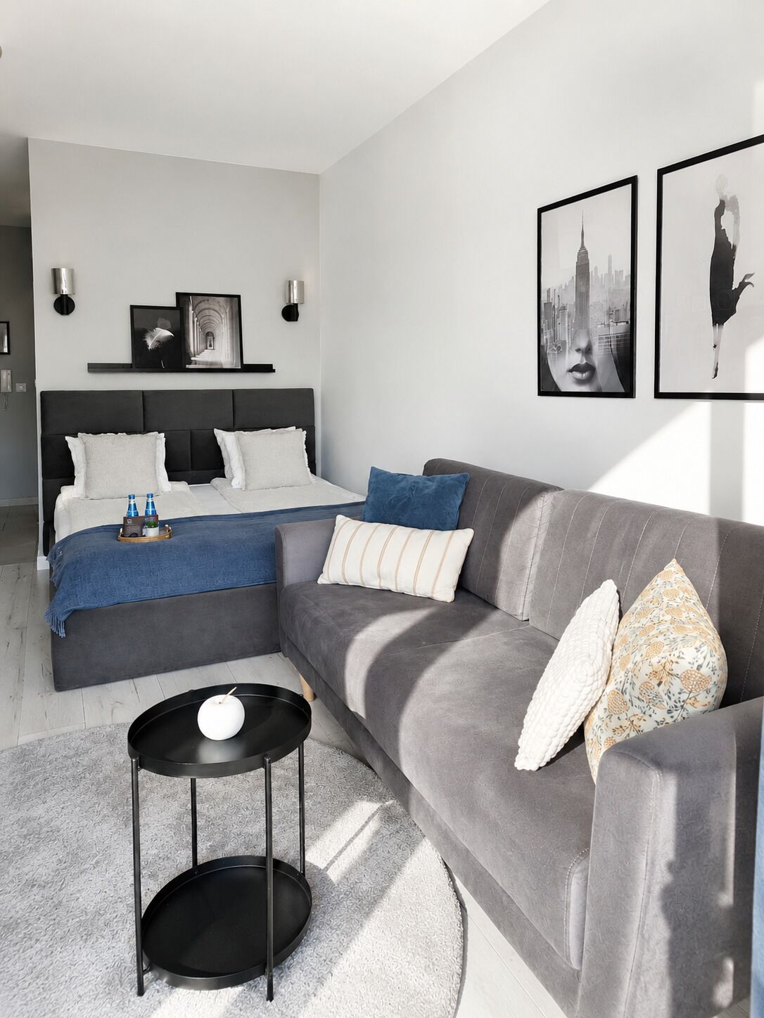 Een moderne slaapkamer met een bed, een grijze bank, een klein rond tafeltje en kunstwerken aan de muur.