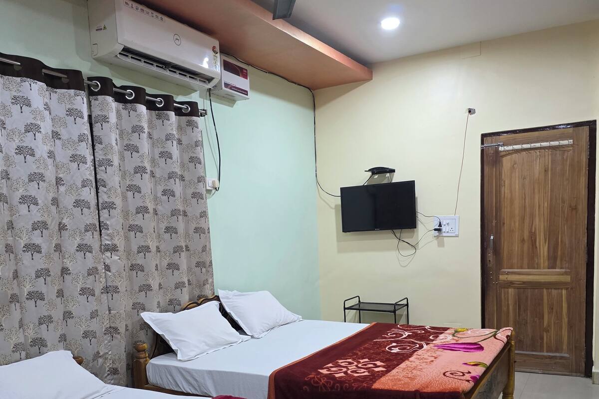 A hotel room with two beds, a ceiling fan, an air conditioning unit, a television mounted on the wall, and a wooden door.