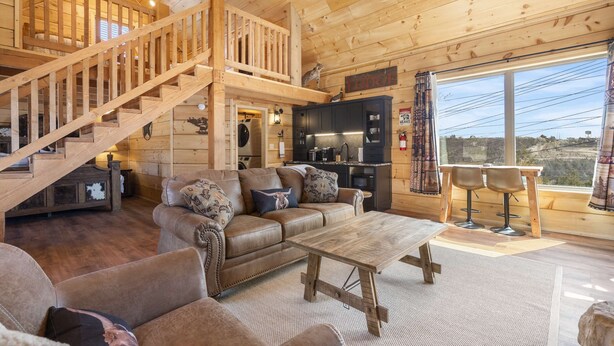 A wooden cabin interior with a staircase, a living room with a sofa and coffee table, a kitchen area, and a large window offering a view. - Elk Lodge – Cabin for 4! (Branson)