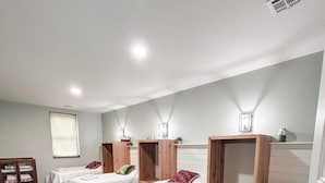 A hotel room with four single beds, each with a wooden headboard and white bedding. There is a window with blinds, a small wooden cabinet, and a wall-mounted shelf with decorative items.