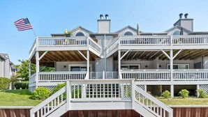 A two-story waterfront house with a balcony, a flag, and a wooden deck. - Sunset Landing: Waterfront Luxury Condo with High End Finishes Throughout (Douglas)