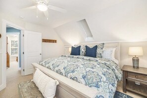 Een slaapkamer met een schuine plafond, een bed met beddengoed met een patroon, een nachtkastje met een lamp en een klein houten nachtkastje.