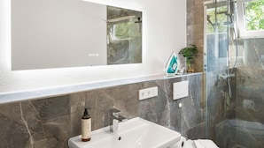 A bathroom with a white sink, a mirror, a shower, and a marble-tiled wall. - Lit Living Deluxe Apartments Wörth (Woerth am Rhein)