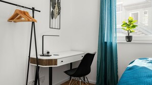 A bedroom with a white desk, black chair, and a bed with blue bedding. There is a window with a potted plant on the windowsill. - Lit Living Deluxe Apartments Wörth (Woerth am Rhein)