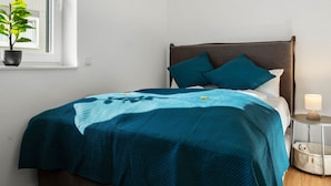 A neatly made bed with a teal blanket and pillows, a bedside table with a lamp, and a potted plant by the window. - Lit Living Deluxe Apartments Wörth (Woerth am Rhein)