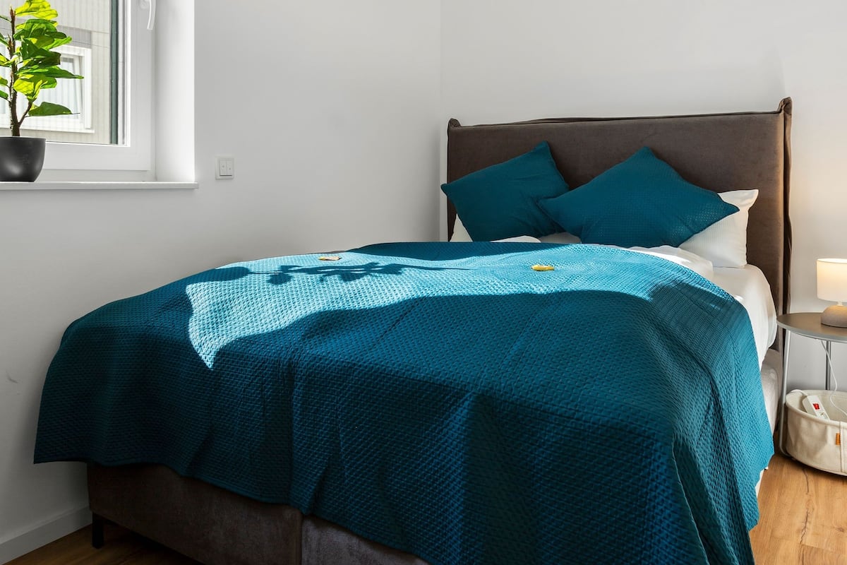 A neatly made bed with a teal blanket and pillows, a bedside table with a lamp, and a potted plant by the window.