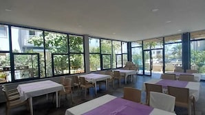 A spacious indoor area with multiple tables and chairs, large windows offering views of greenery and buildings outside. - Anamur Çilek Otel (Anamur)
