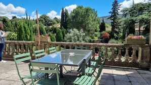 Un patio con mesa de vidrio y sillas verdes, rodeado por una barandilla de piedra y abundante vegetación.