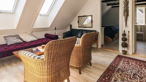Ein Loft-Zimmer mit schrägem Dach, einer Couch, zwei Korbstühlen, einem Spiegel und Blick auf ein Schlafzimmer durch eine offene Tür.