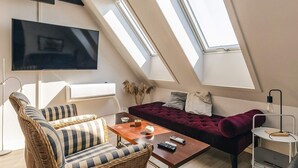 Eine moderne Loft-Wohnung mit schrägem Dach, einem großen Flachbildfernseher, einem Korbssessel, einem roten Samtsofa und einem kleinen Tisch mit Fernbedienungen.