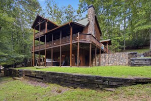 A two-story log cabin with a stone chimney, a covered porch, and a stone retaining wall.