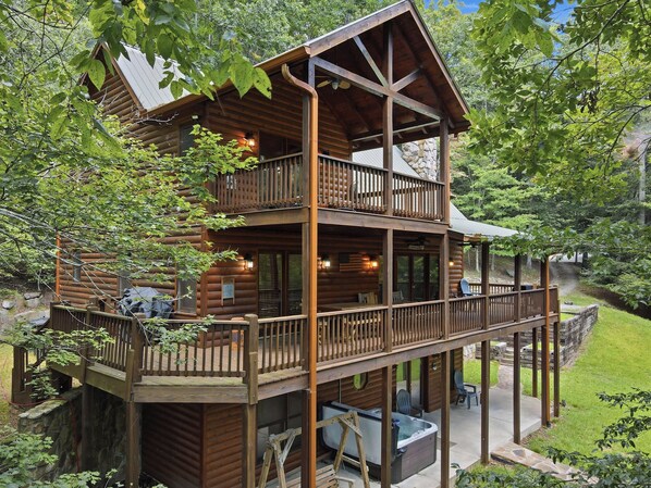 A two-story log cabin with a balcony, hot tub, and outdoor seating area surrounded by trees.