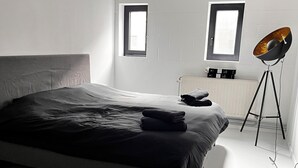 A modern bedroom with a large bed, two windows, a radiator, and a floor lamp. - Discover our unique design loft in downtown Mons (Mons)