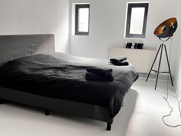 Une chambre moderne avec un grand lit, deux fenêtres, un radiateur et une lampe sur pied.