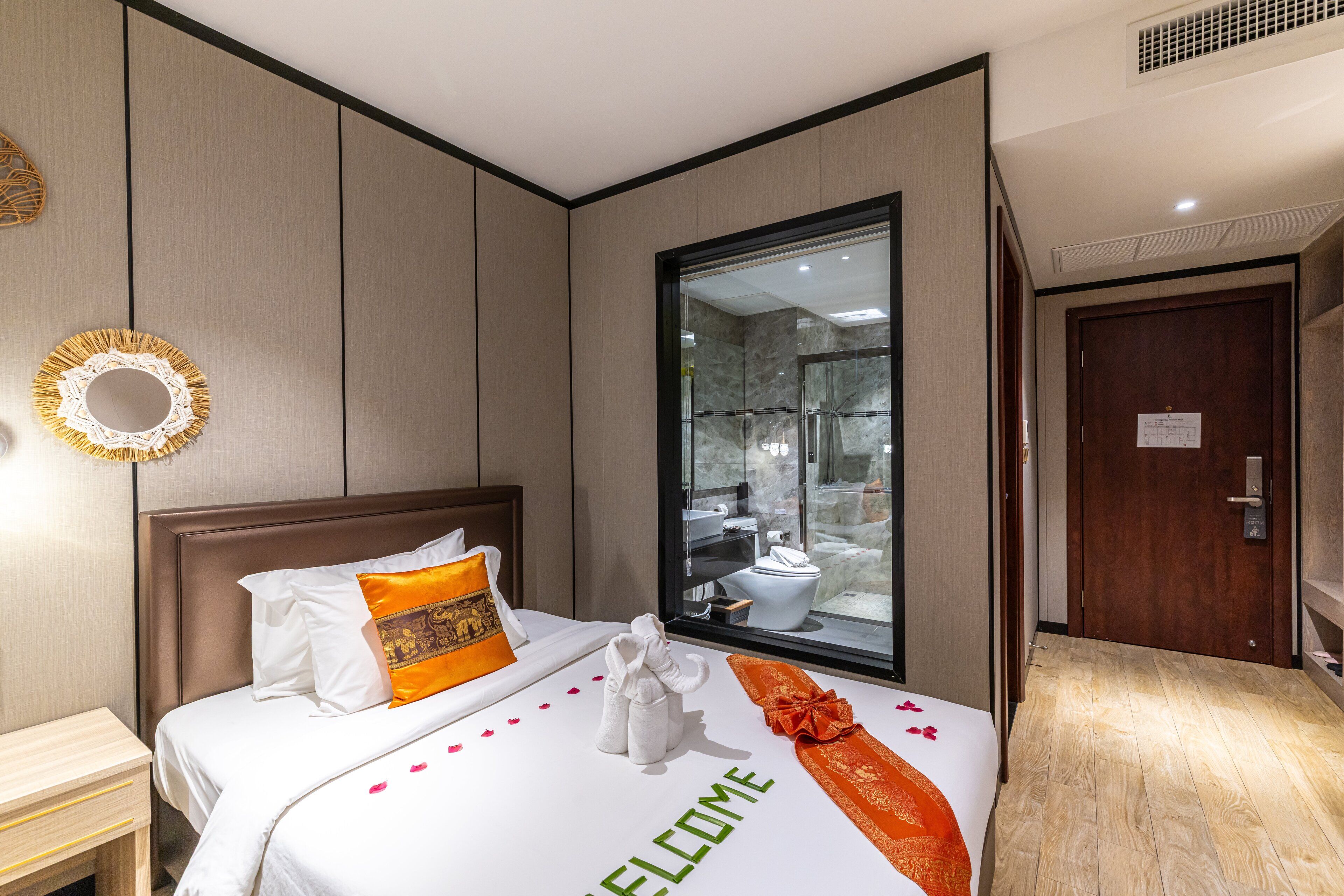 A hotel room with a bed, a wooden nightstand, a mirror, and a view of the bathroom through a glass door.