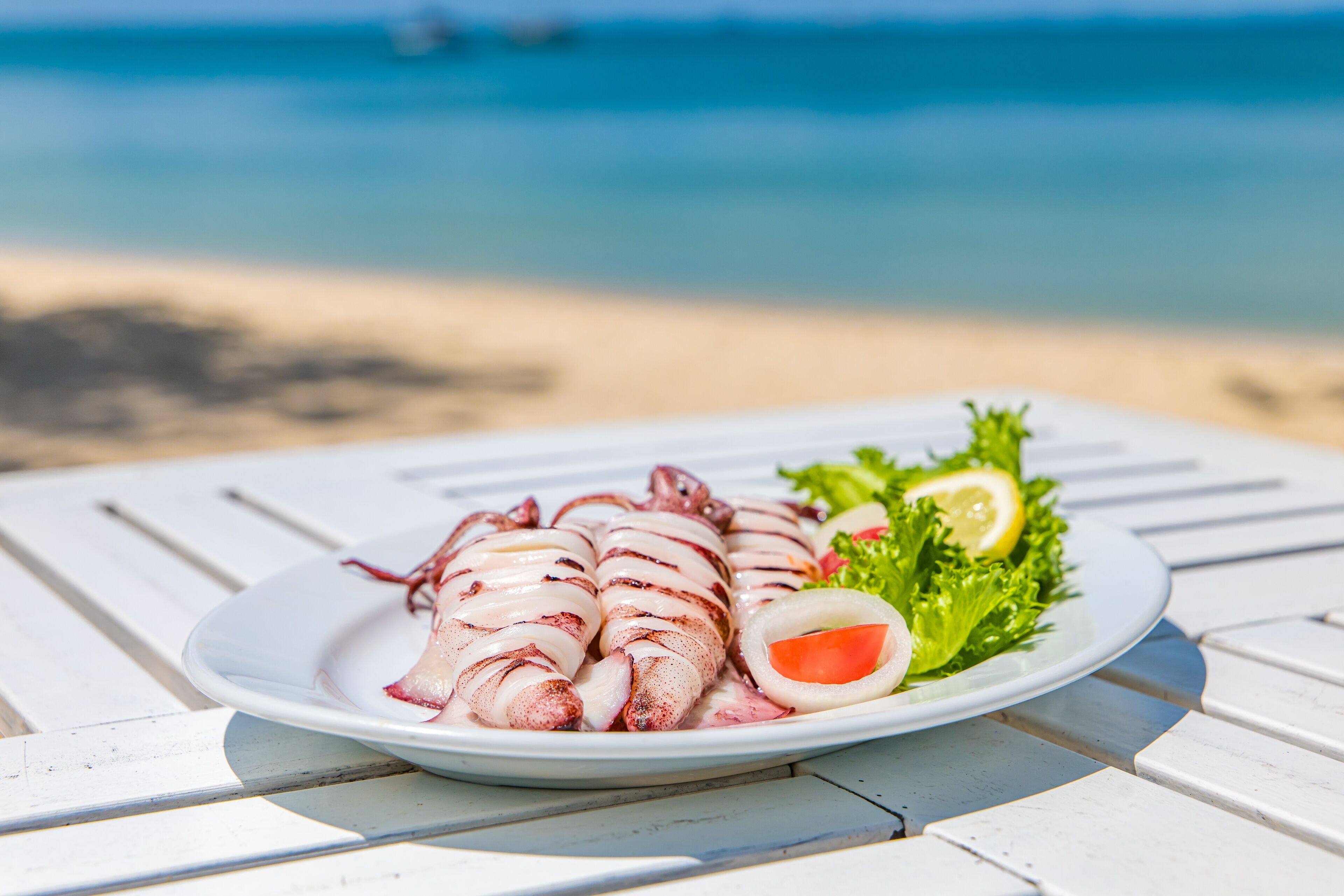 Une assiette de calamars grillés, servis avec du citron et de la laitue, sur une table blanche au bord de la plage.