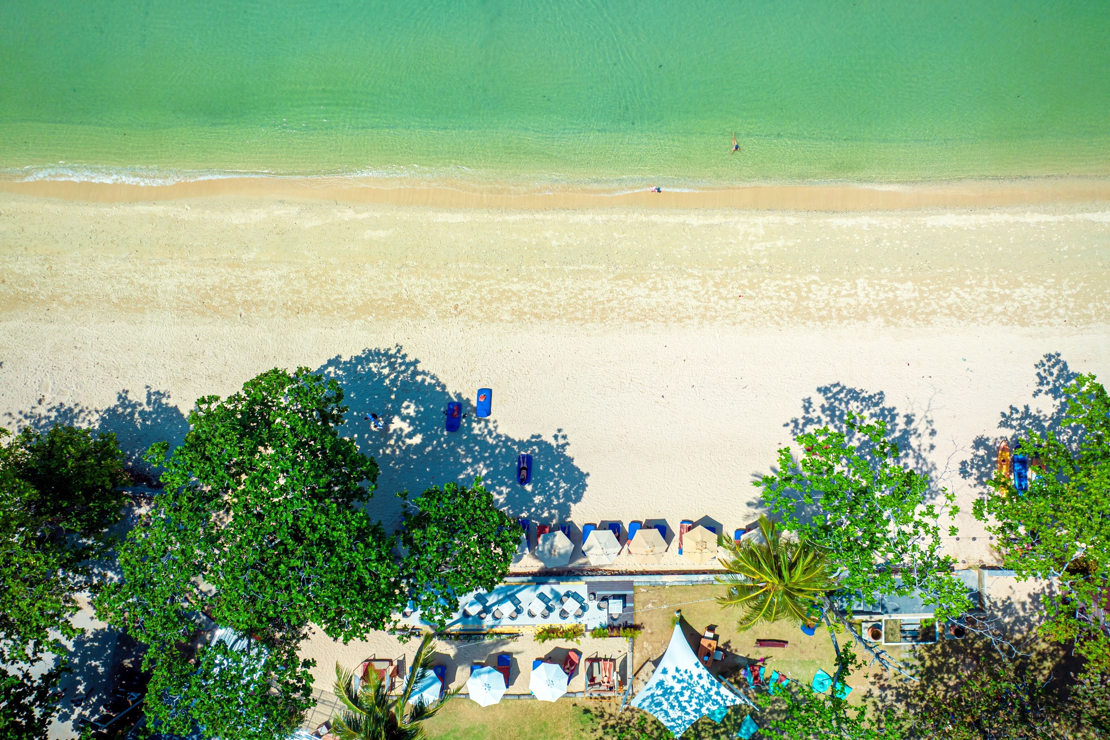 An aerial view of a beach with clear turquoise water, a sandy shore, and a row of blue umbrellas and lounge chairs.