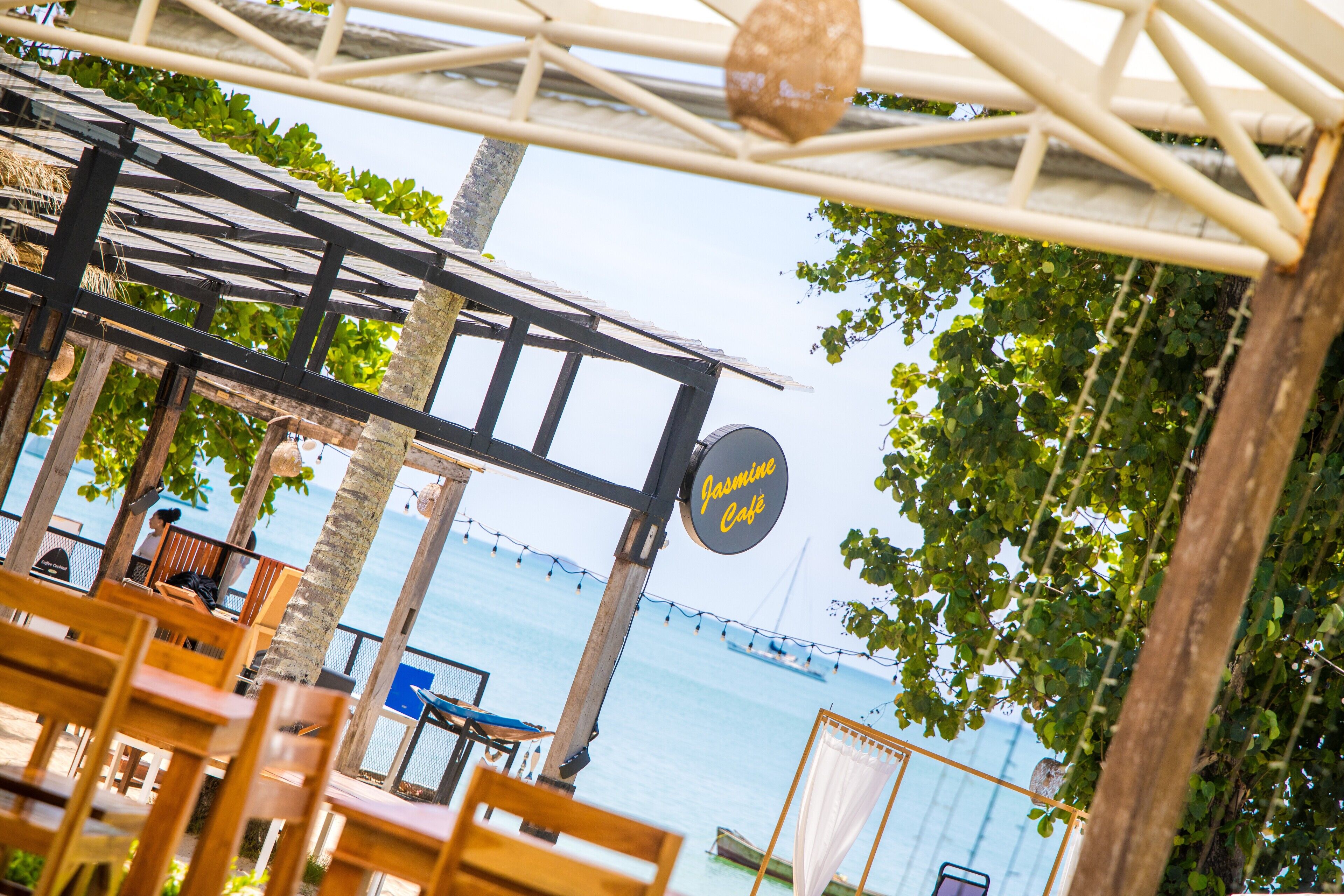 Ein Straßencafé mit Holztischen und -stühlen, einem Schild mit der Aufschrift "Jasmine Café" und Blick auf das Meer mit Booten im Hintergrund.