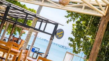 Ein Straßencafé mit Holztischen und -stühlen, einem Schild mit der Aufschrift "Jasmine Café" und Blick auf das Meer mit Booten im Hintergrund.