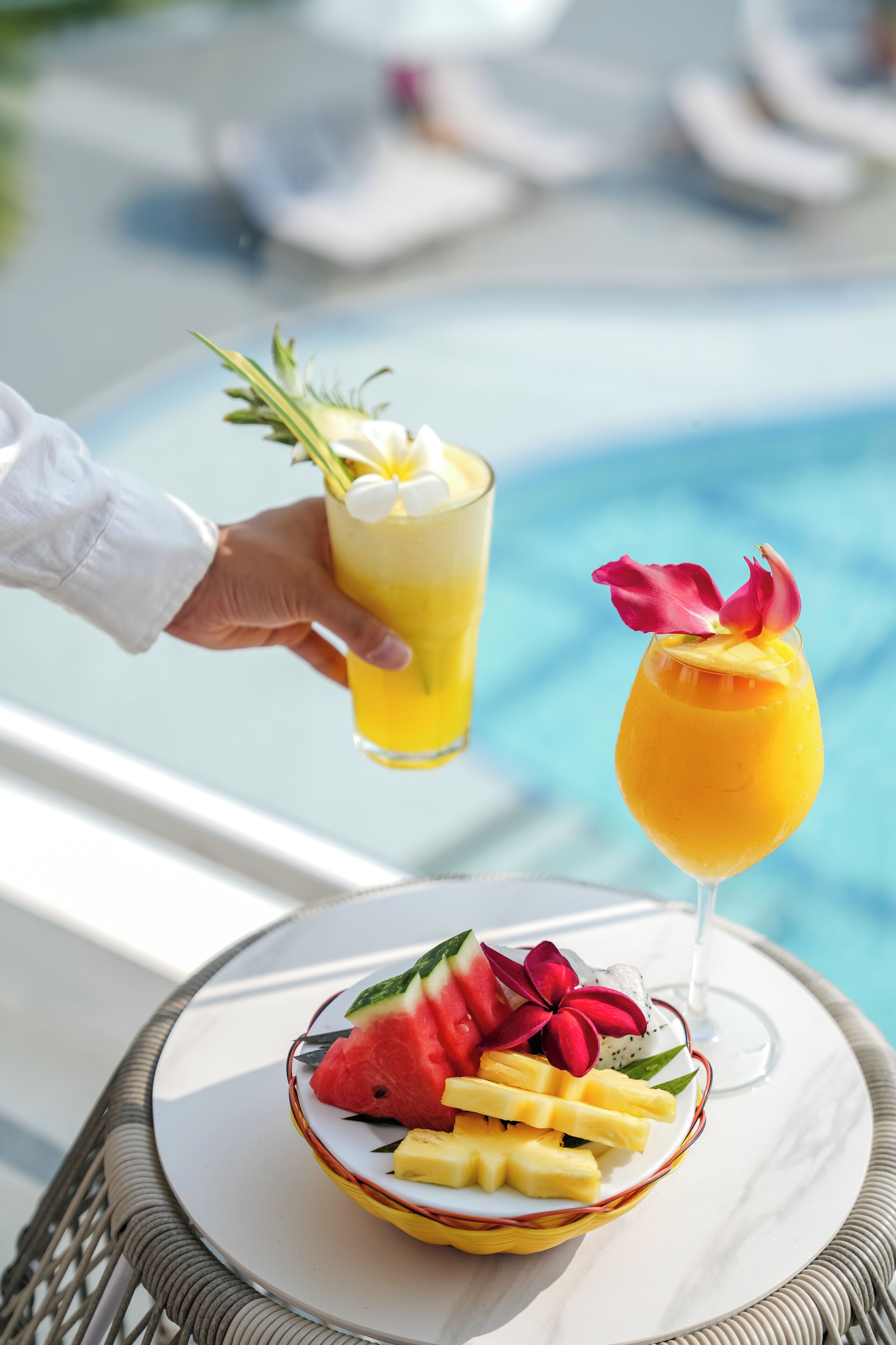 A table with a fruit bowl and two drinks, one with a flower garnish, near a pool.