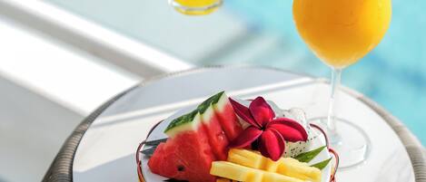 A table with a fruit bowl and two drinks, one with a flower garnish, near a pool.