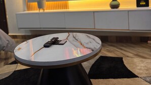 Un salón moderno con una mesa redonda de mármol, un televisor de pantalla plana montado en la pared, una planta en maceta y un mueble blanco debajo.