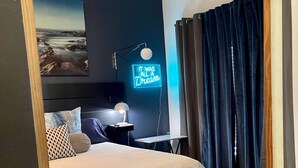 Une chambre avec un lit, un bureau, une chaise, un ventilateur de plafond, un néon et une vue sur l’extérieur.