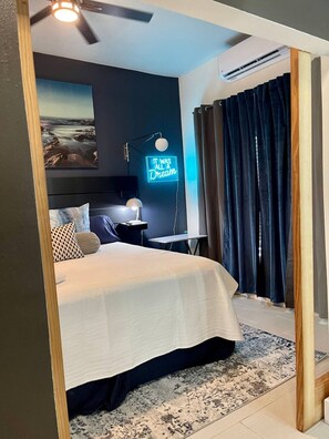 A bedroom with a bed, a desk, a chair, a ceiling fan, a neon sign, and a view of the outdoors. - Coastal Retreat Near Pleasure Pier & Beach (Galveston)