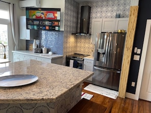 A modern kitchen with a granite countertop, stainless steel appliances, a patterned backsplash, and a flat-screen TV mounted above the stove. - Coastal Retreat Near Pleasure Pier & Beach (Galveston)