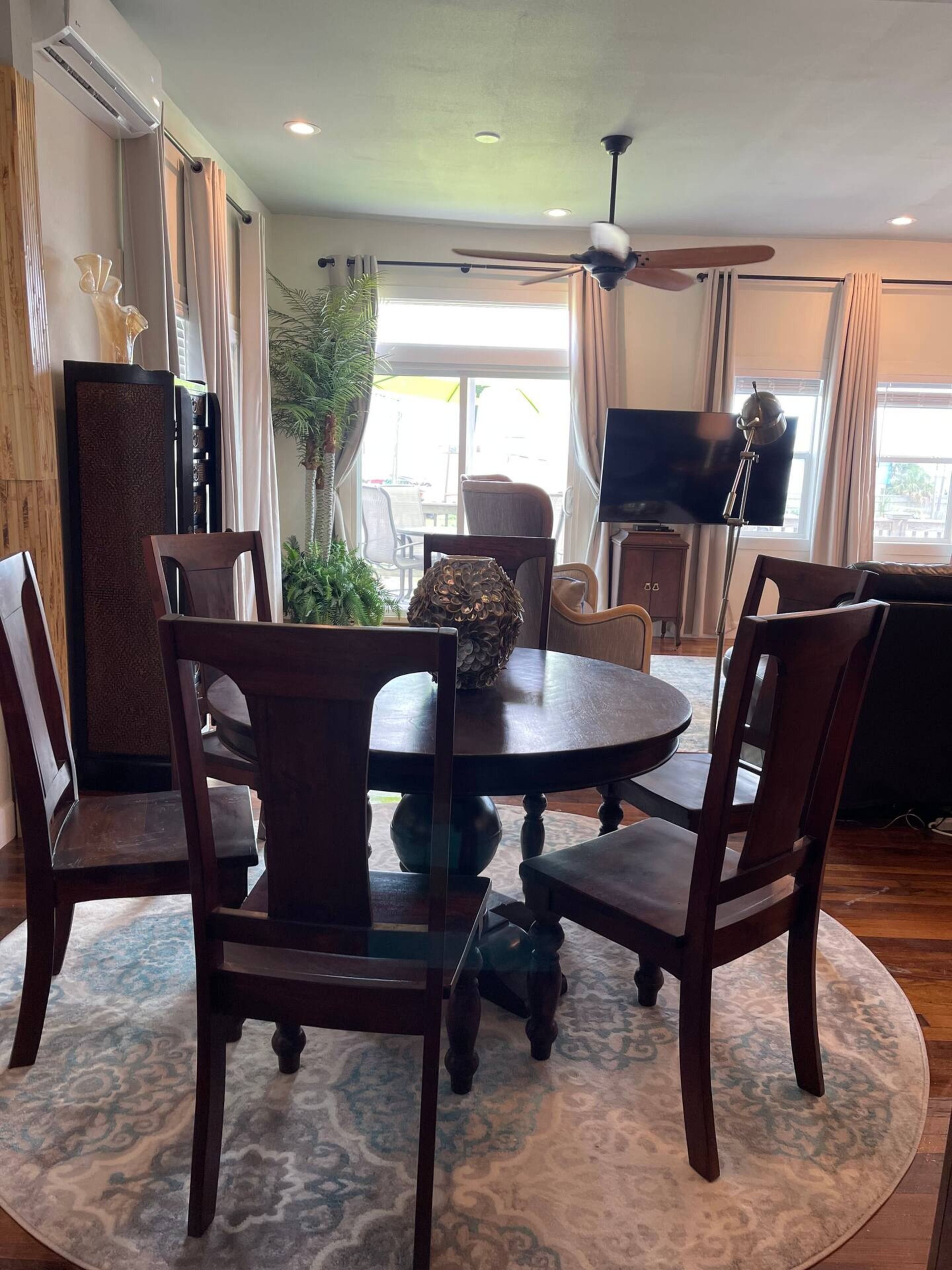 A dining area with a round table, six chairs, a ceiling fan, a TV, and a large window with curtains.