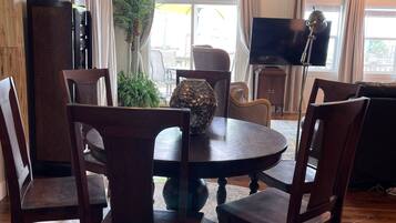 A dining area with a round table, six chairs, a ceiling fan, a TV, and a large window with curtains.